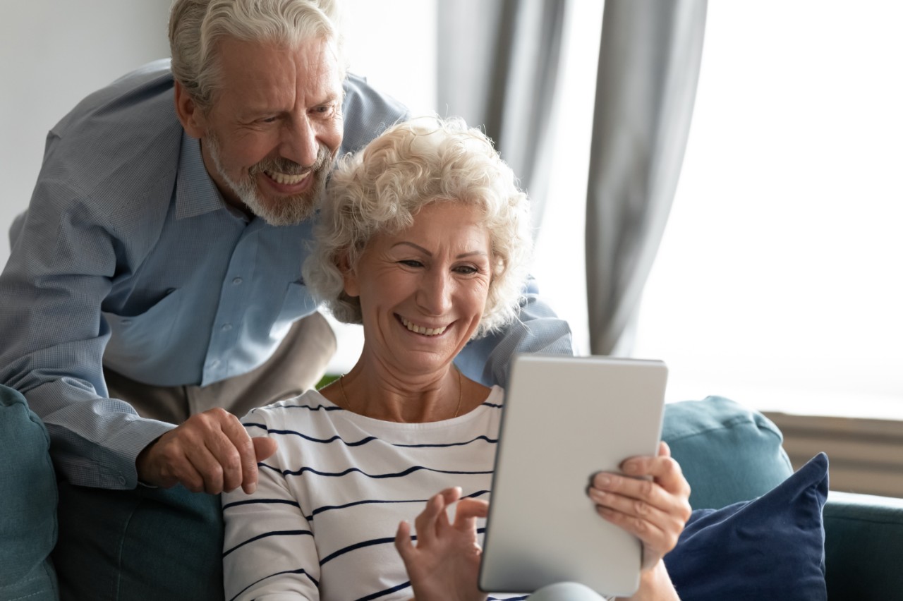 Coppia anziana seduta sul divano, guardando un iPad, ridendo e sorridendosi reciprocamente