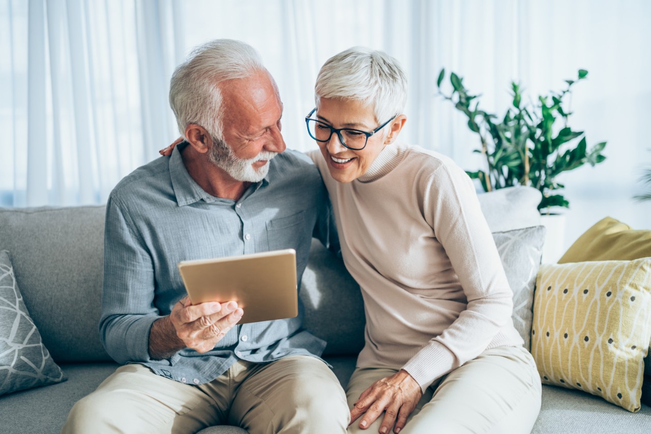 Old couple sitting on their sofa, looking at an iPad, laughing and smiling at each other