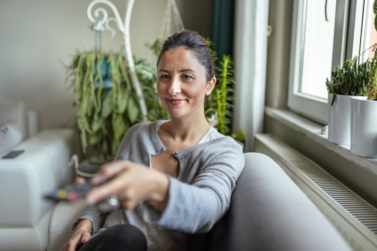  Eine junge Frau hält eine Fernbedienung, während sie Hörgeräte auf ihrem Sofa benutzt